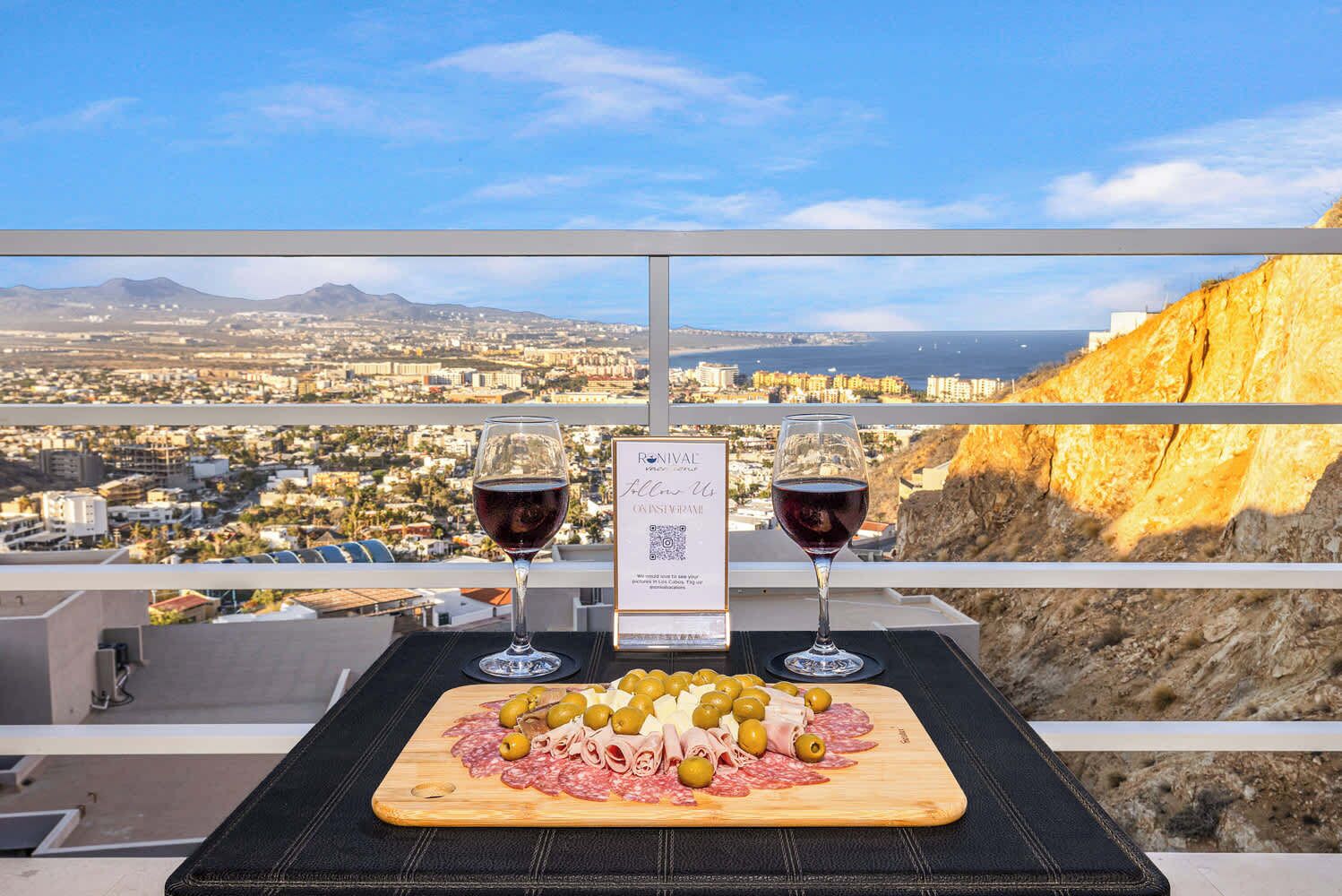 A table with two wine glasses, a menu, and a charcuterie board with olives and meats, overlooking a cityscape and mountains.
