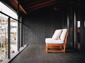 A modern outdoor seating area with a wooden chair and cushions, a dark floor, and a view through large windows.