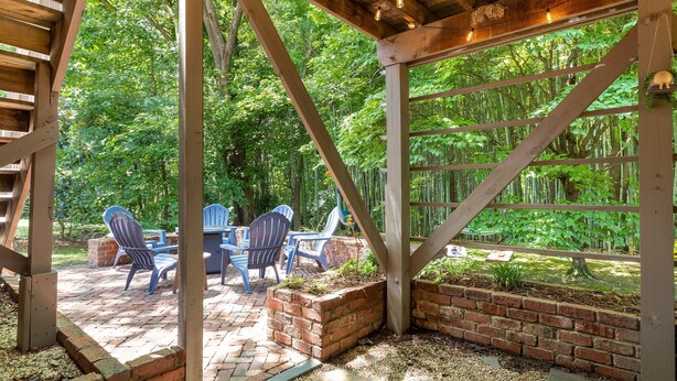 A covered patio with brick planters and blue chairs, surrounded by lush greenery. - Fenced Yard | Pool Access | Fire Pit (Wake Forest)