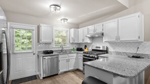 A modern kitchen with white cabinets, a granite countertop, stainless steel appliances, and a window with blinds. - Fenced Yard | Pool Access | Fire Pit (Wake Forest)