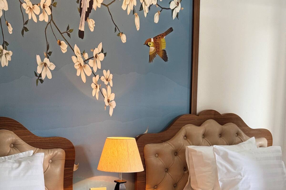 A hotel room with two beds, a nightstand with a lamp, and a wall with floral wallpaper.