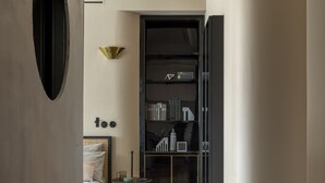A modern bedroom with a wooden parquet floor, a bed with a headboard, a bedside table, and a wall-mounted shelf with books and decorative items. - Homy® - The Signature Suite 2 (Milan)