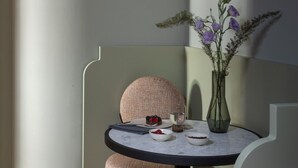 A modern, compact room with a round table, a cushioned chair, a vase with flowers, and a small dessert on the table.