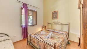 Un dormitorio con cama, ventana con cortinas, mesita de noche y un cuadro colgado en la pared.