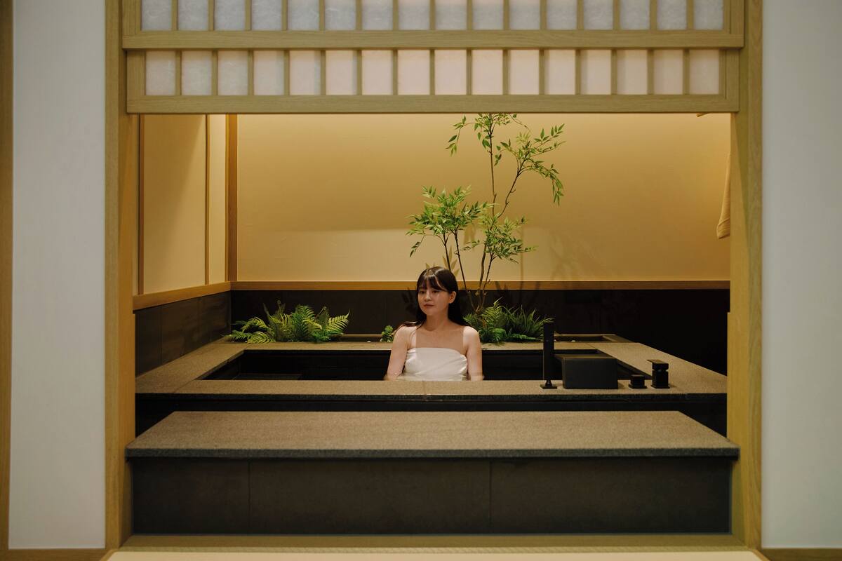 A woman sitting at a counter with a plant and a small table in a room with wooden paneling and a low ceiling.