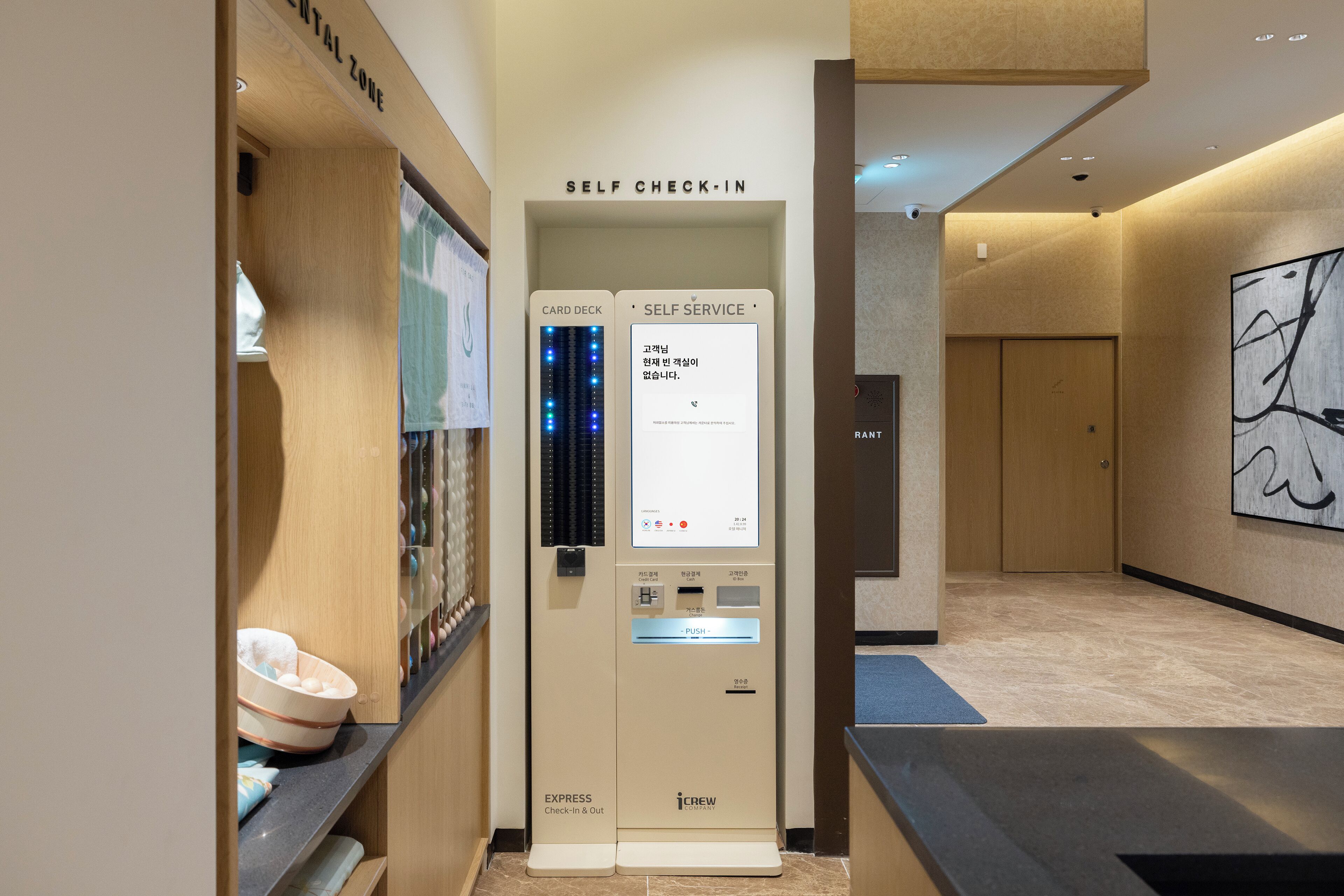 A self-service check-in kiosk with a card deck and self-service options, located in a modern hotel lobby with a minimalist design and a large abstract painting on the wall.