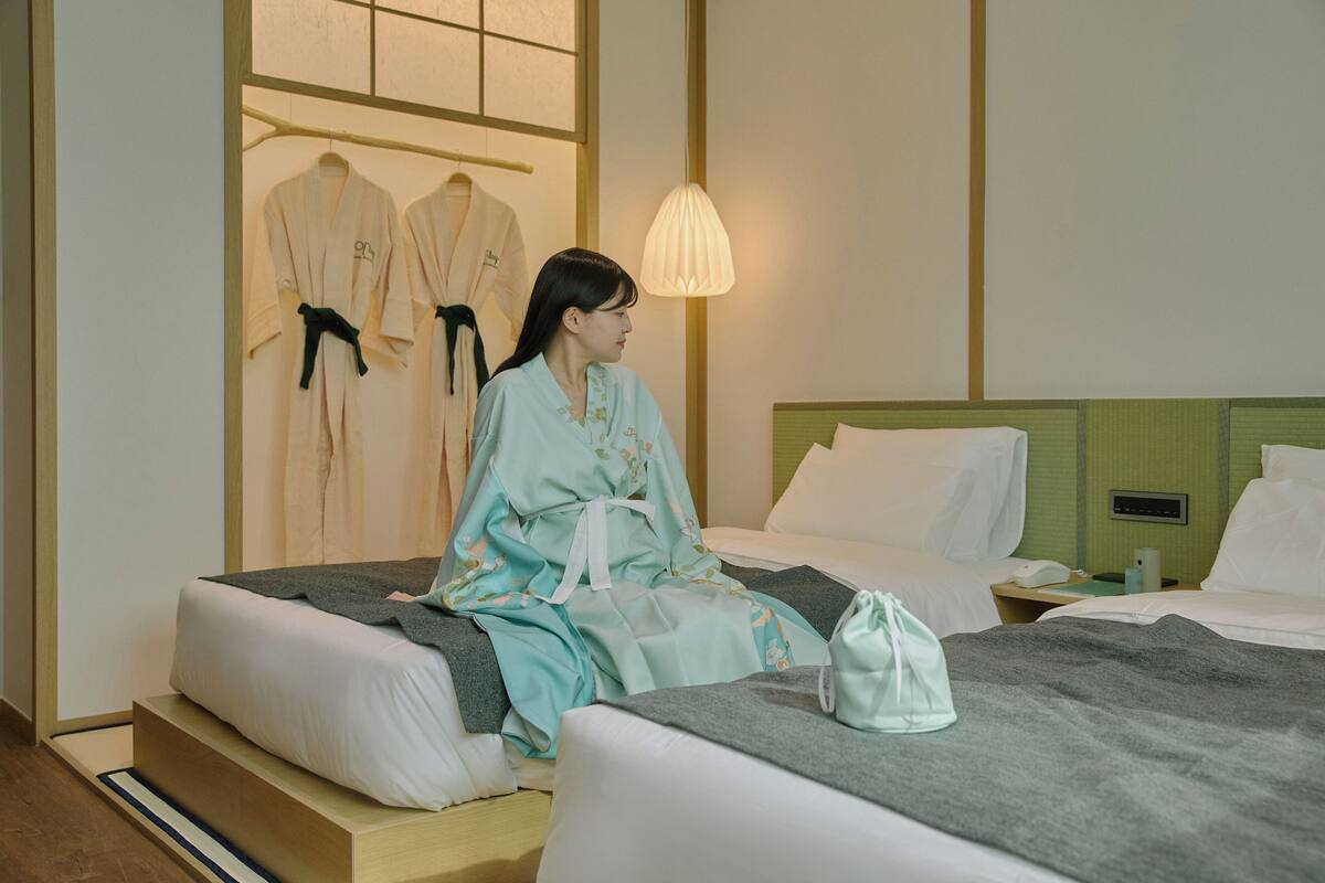 A hotel room with two beds, a bedside table with a lamp, and a wall-mounted TV.