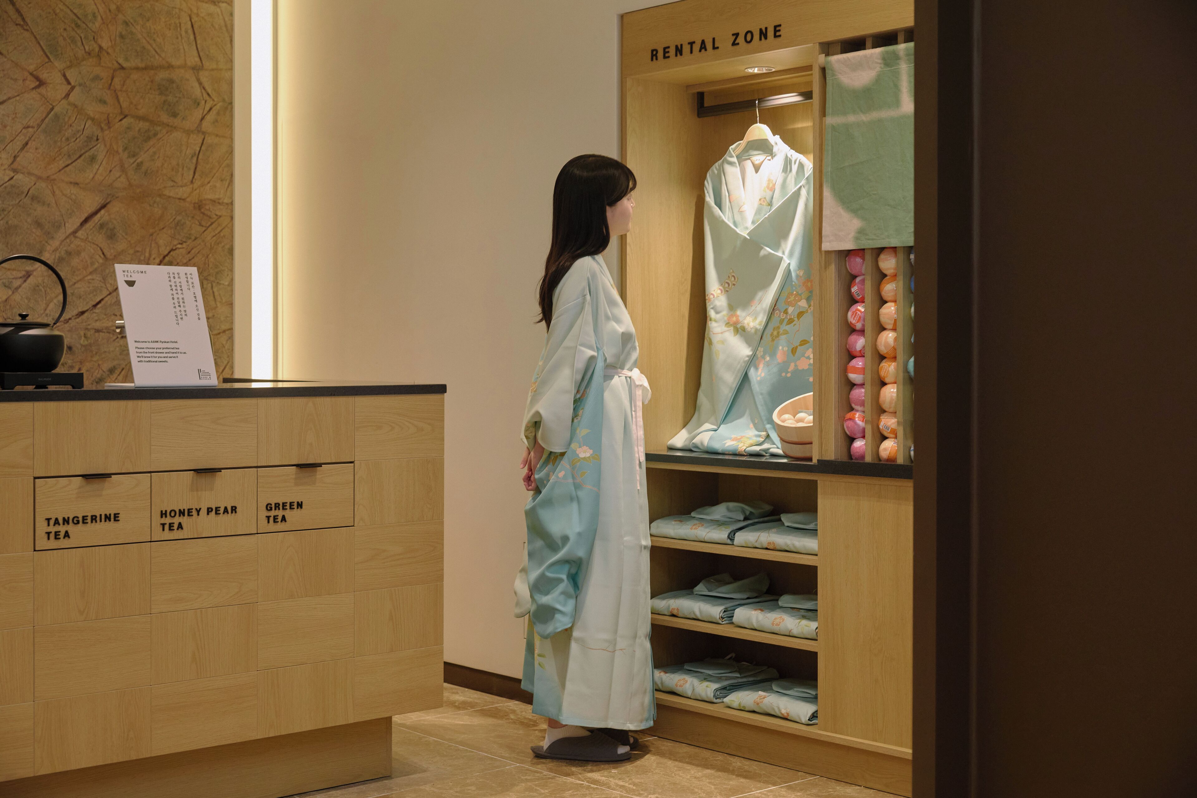 A woman in a robe standing in a room with a wooden counter labeled 'RENTAL ZONE' and shelves with folded robes.