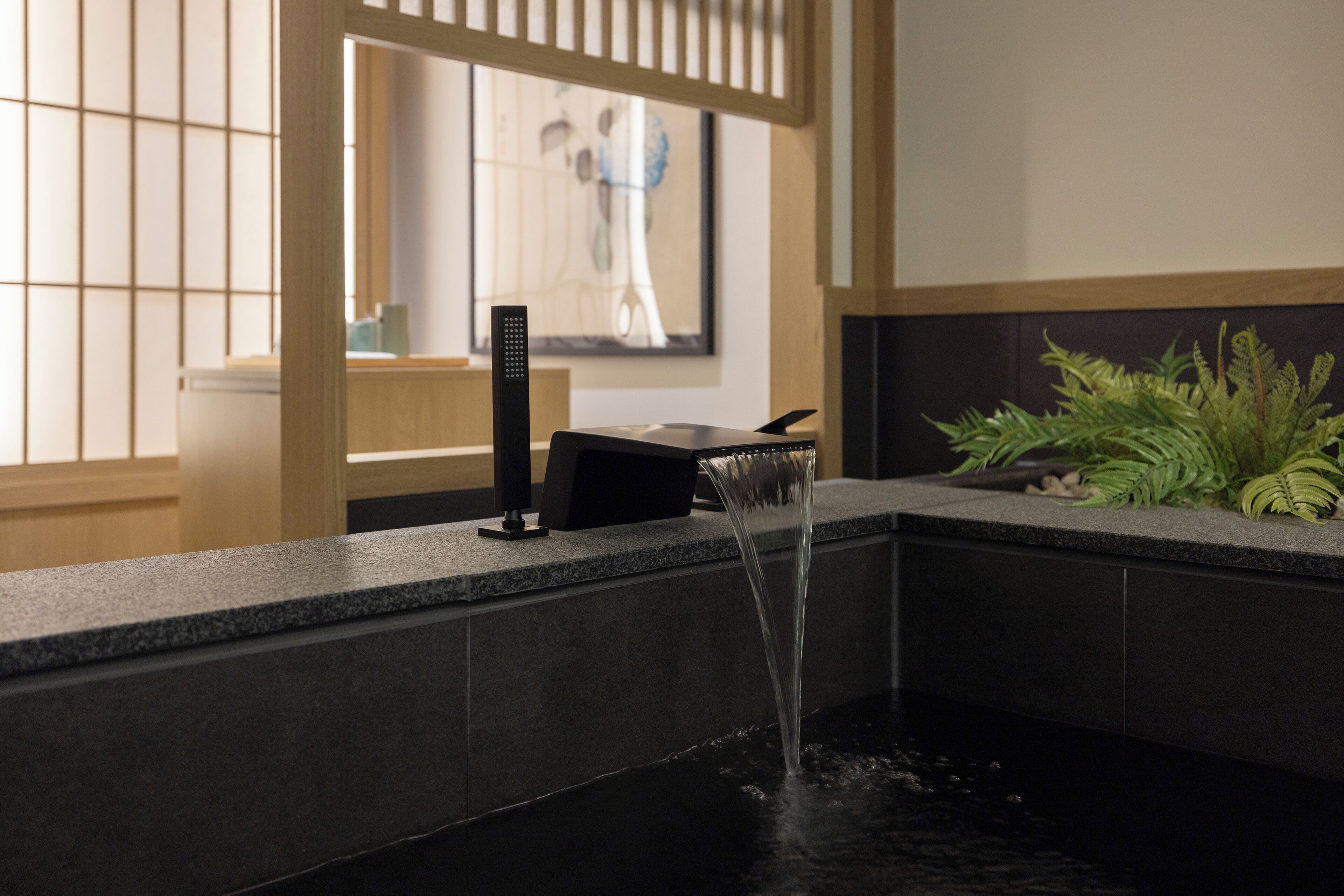 Ein modernes Badezimmer mit Wasserspiel, einem schwarzen Wasserhahn und einer Topfpflanze.