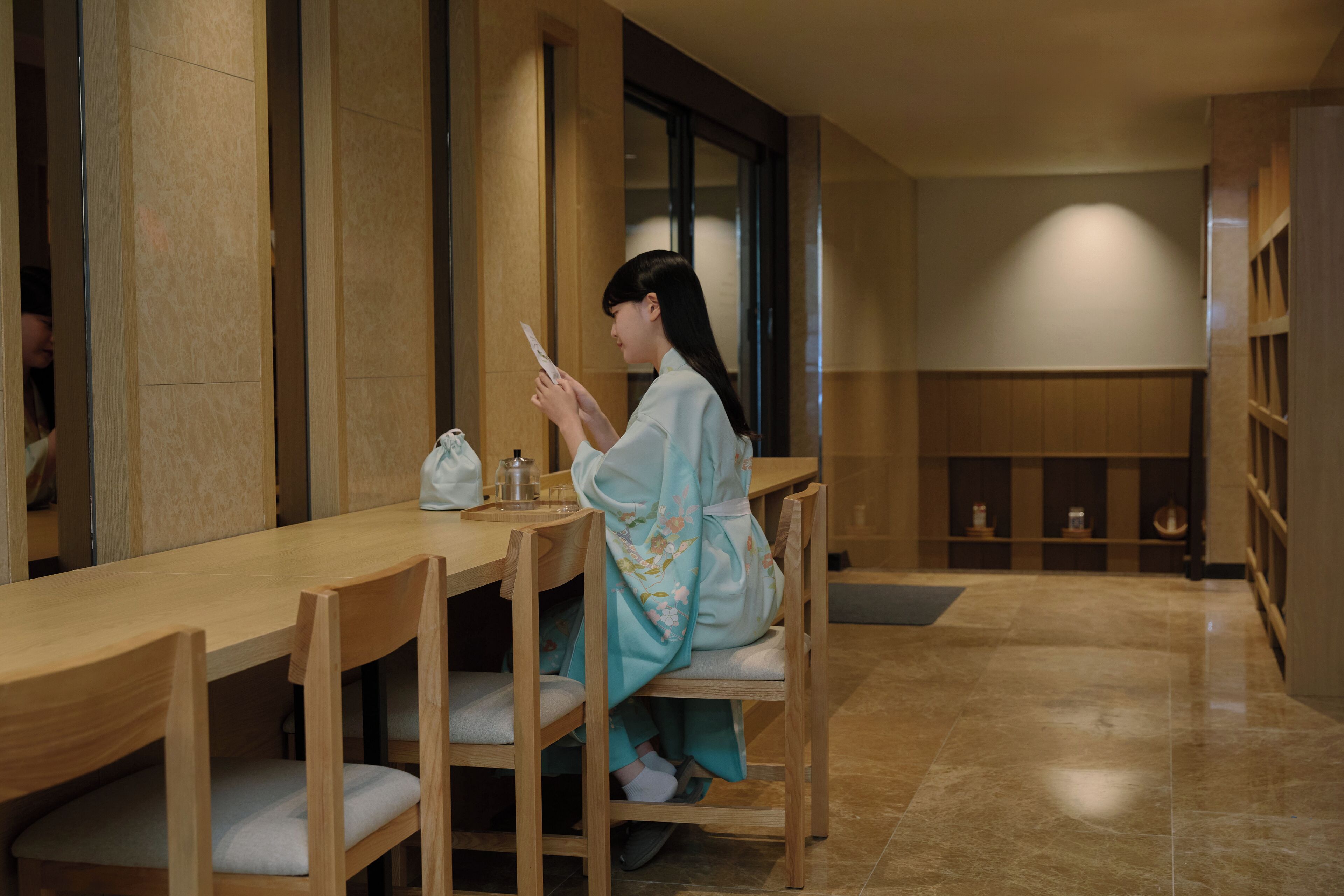 A woman sitting at a wooden table in a room with a tiled floor and wooden shelves.