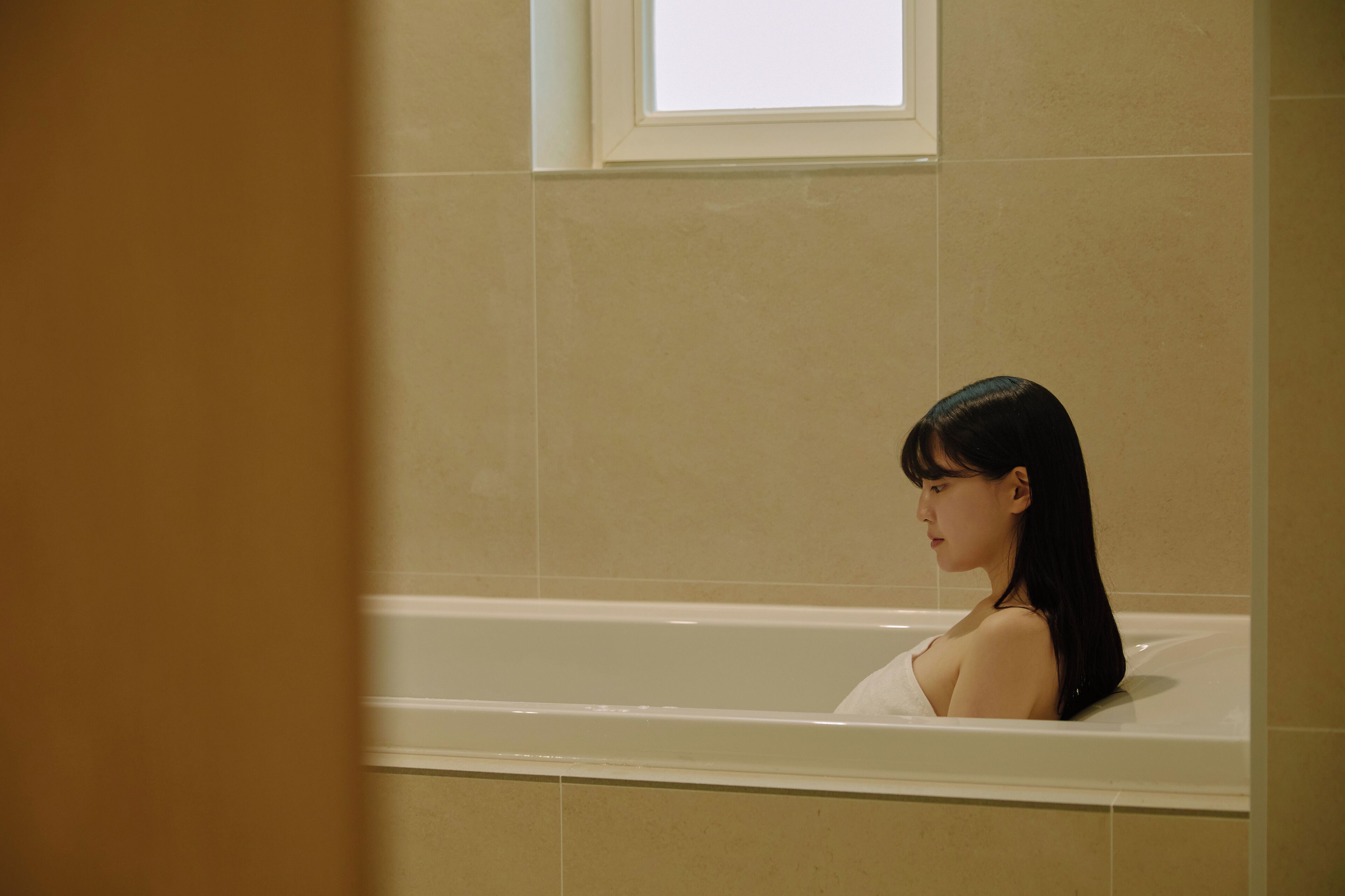 A woman in a bathtub in a room with beige tiles and a window above the tub.