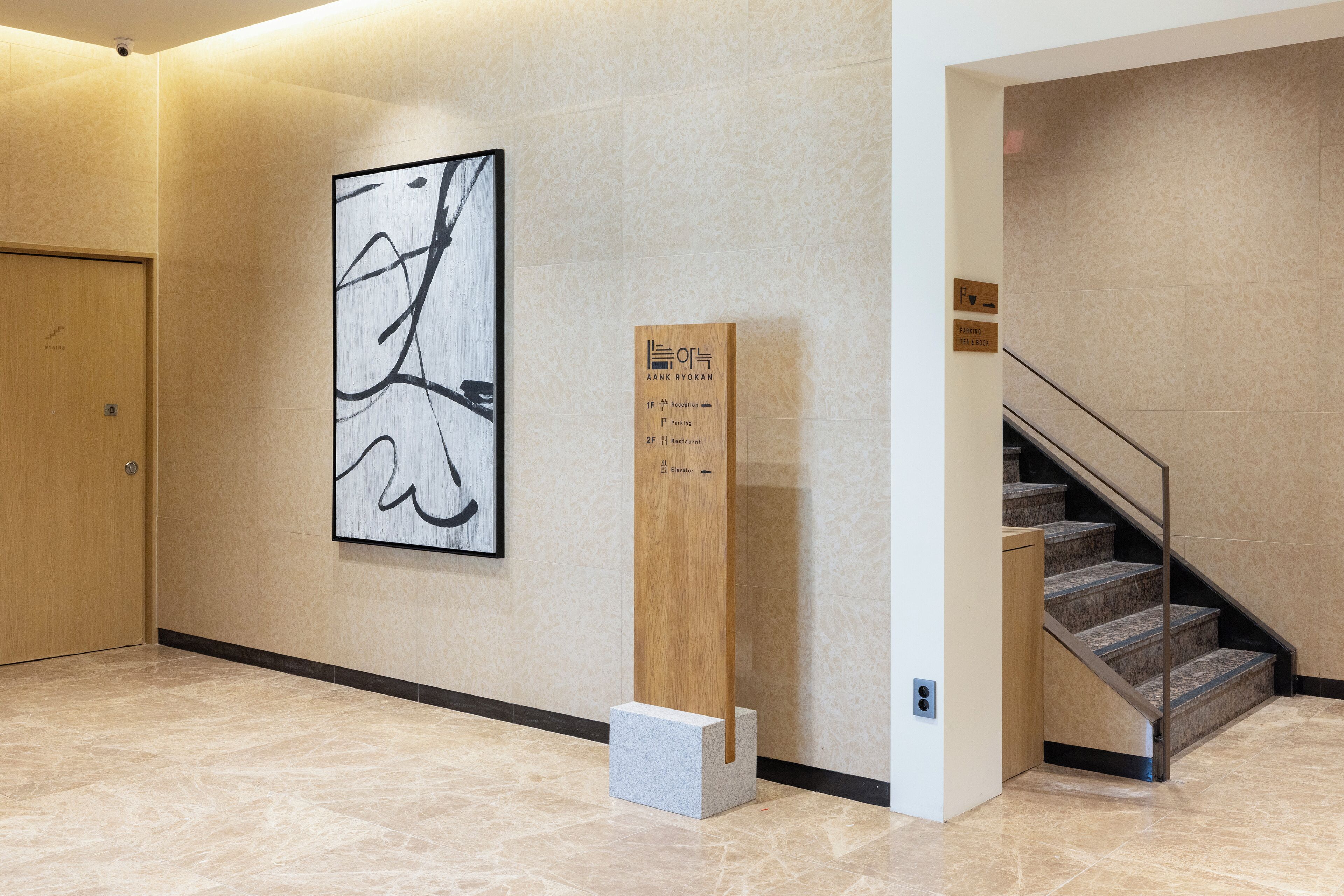 Un vestíbul d'hotel amb sòl de marbre, una escala, un senyal de fusta i una obra d'art abstracta a la paret.