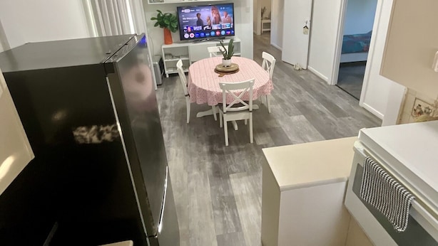 Cocina compacta con refrigerador, horno y microondas. Hay un comedor con mesa y sillas, y un televisor al fondo.
