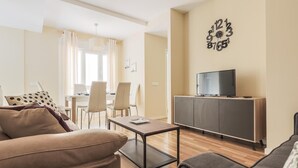 Una sala de estar moderna con un sofá, una mesa de centro, un televisor sobre un mueble bajo y una zona de comedor con sillas y una mesa.