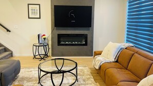 A modern living room with a flat-screen TV mounted on a grey wall, a fireplace below it, a brown leather sofa, a glass coffee table, and a small side table with a vase of flowers.