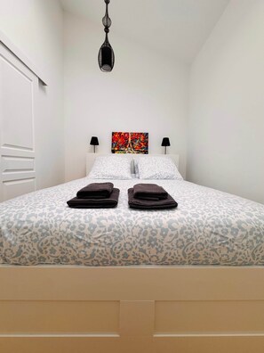 A bedroom with a bed, two pillows, two folded towels, a painting of the Eiffel Tower, and a hanging light fixture.
