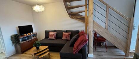 A modern living room with a wooden staircase, a black sofa, a wooden dining table, and a television.