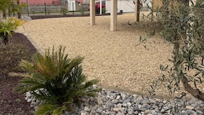 Um bairro residencial com uma casa moderna, uma entrada com calçamento de brita e um pequeno jardim com pedras e plantas.