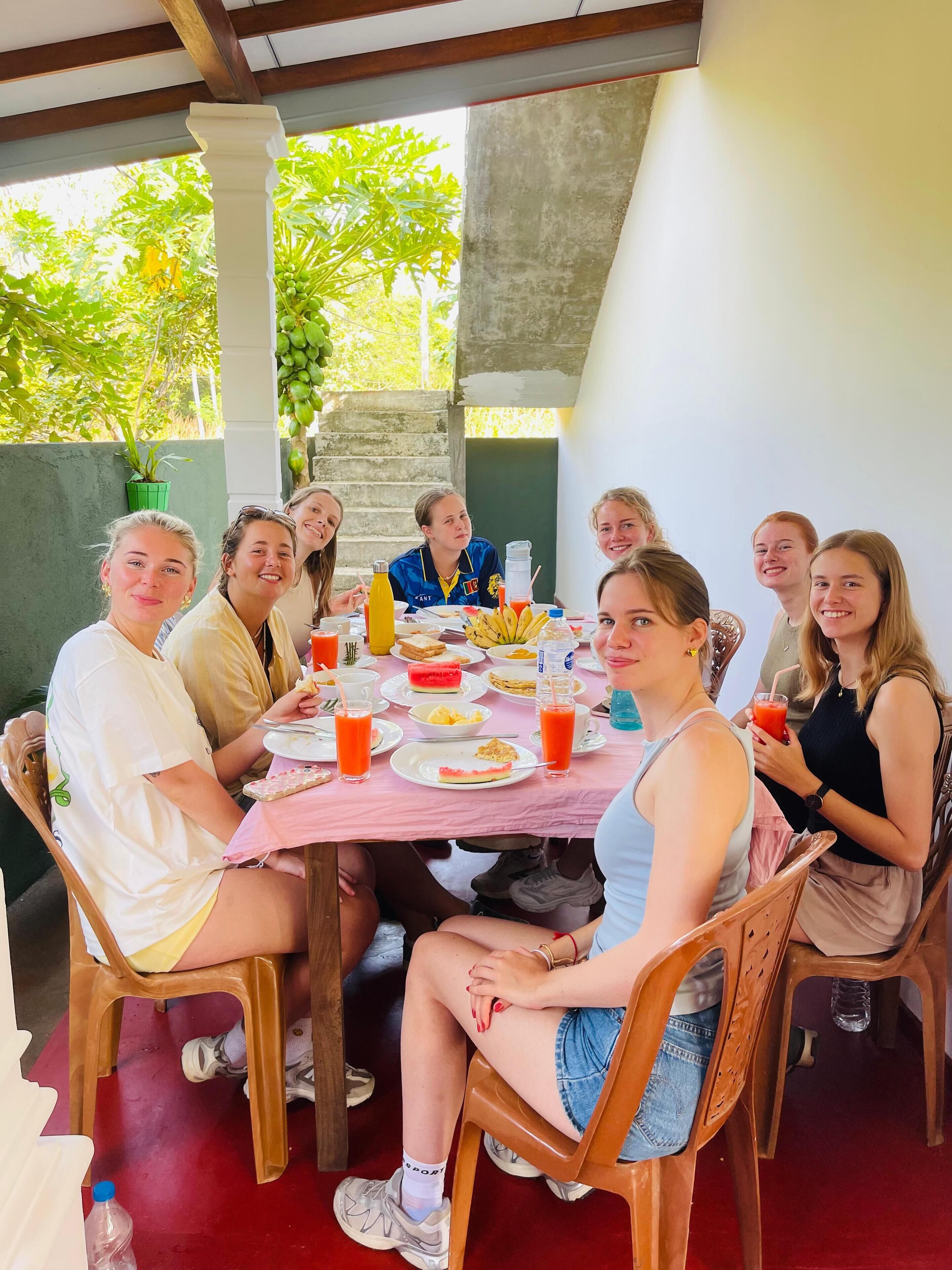 Un gruppo di persone sedute a tavola con cibo e bevande, in un ambiente esterno con una struttura in cemento e vegetazione sullo sfondo.