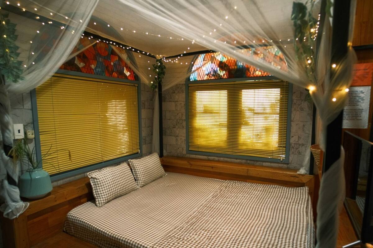 Una habitación acogedora con una cama a cuadros, ventanas amplias con persianas y luces colgantes.
