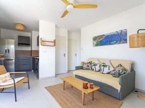 Un salon moderne avec un canapé, une table basse, un fauteuil en osier, un ventilateur de plafond et une cuisine en arrière-plan.