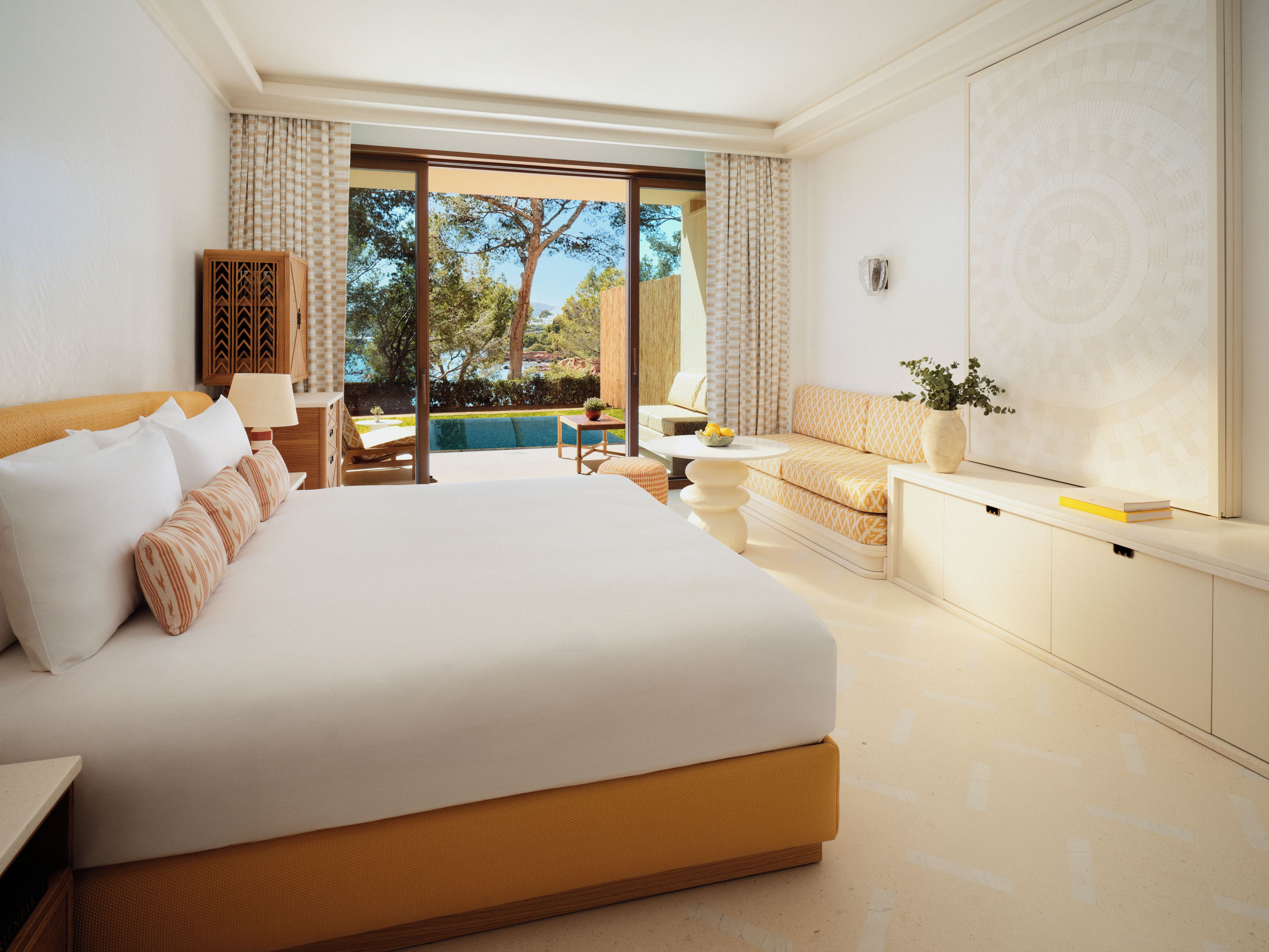 A hotel room with a large bed, a small table, a sofa, and a view of a pool and trees.