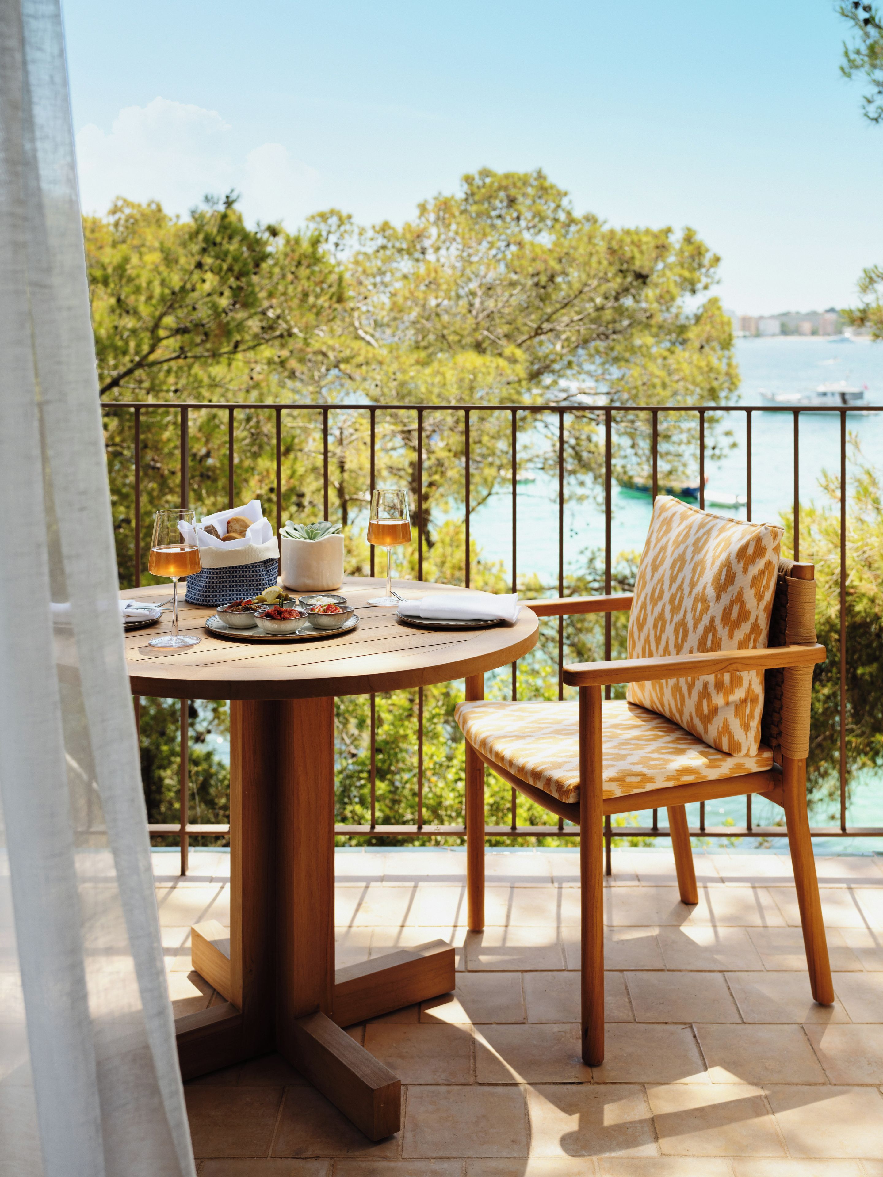 Une table ronde en bois, accompagnée d’une chaise, d’un verre et d’un panier de pain, est placée sur un balcon doté d’une balustrade.