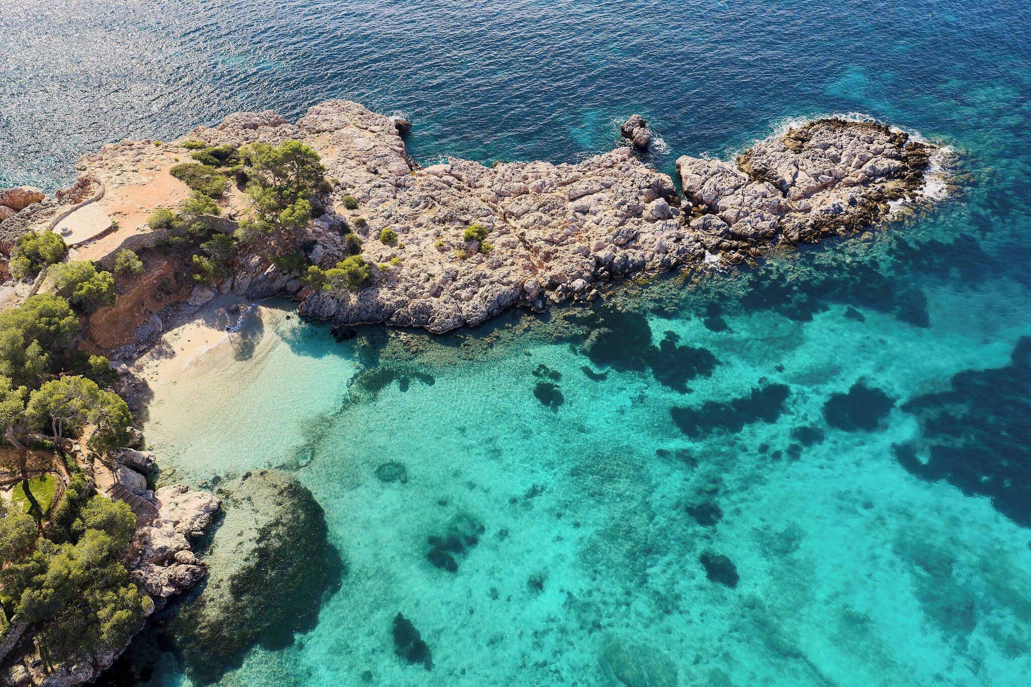 Vista aérea de una costa rocosa con aguas turquesas cristalinas, una playa de arena y árboles verdes frondosos.