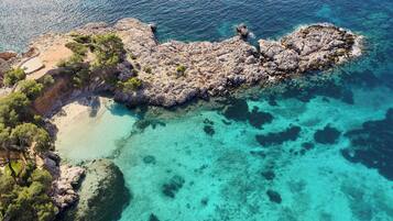 Vista aérea de una costa rocosa con aguas turquesas cristalinas, una playa de arena y árboles verdes frondosos.