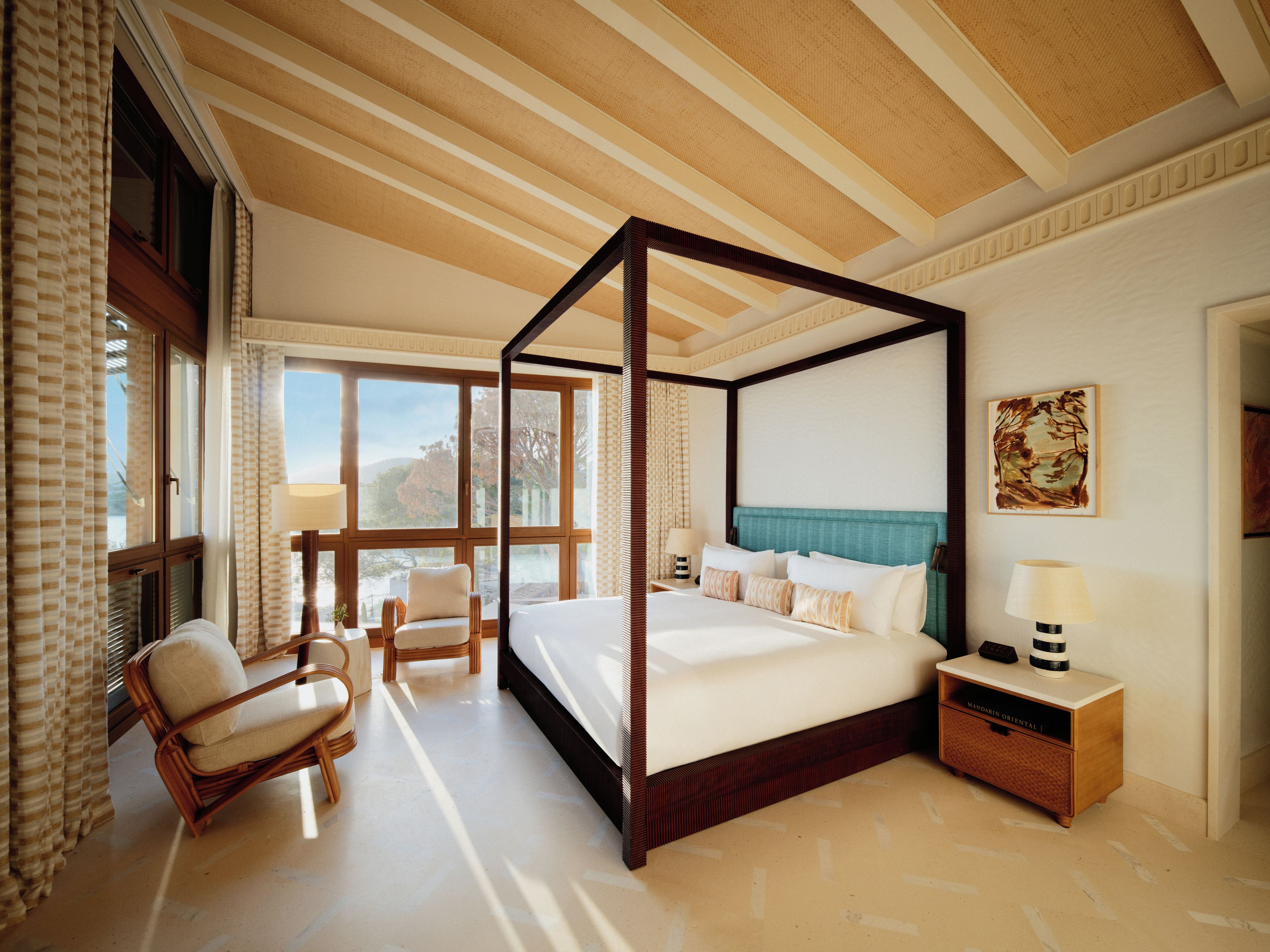 Una cama con dosel con estructura de madera, una mesita de noche con lámpara y una silla en una habitación con ventanas grandes y vistas al exterior.