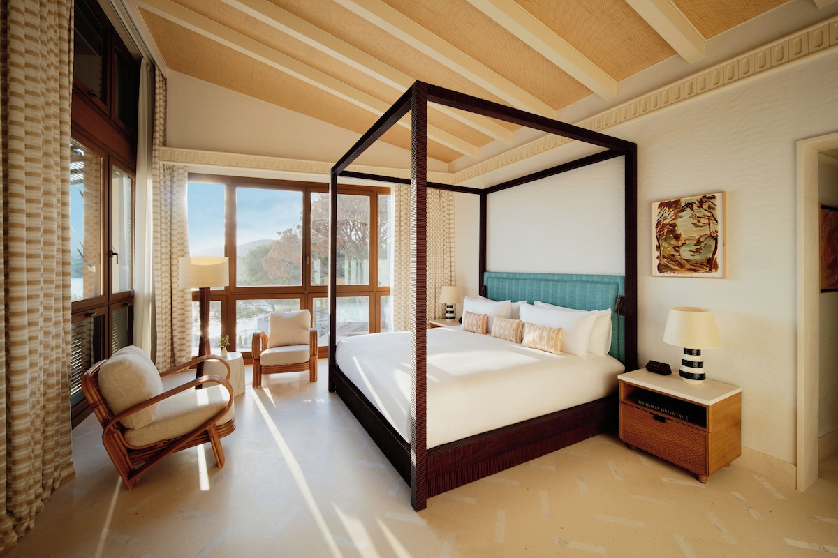 A four-poster bed with a wooden frame, a bedside table with a lamp, and a chair in a room with large windows and a view of the outdoors.