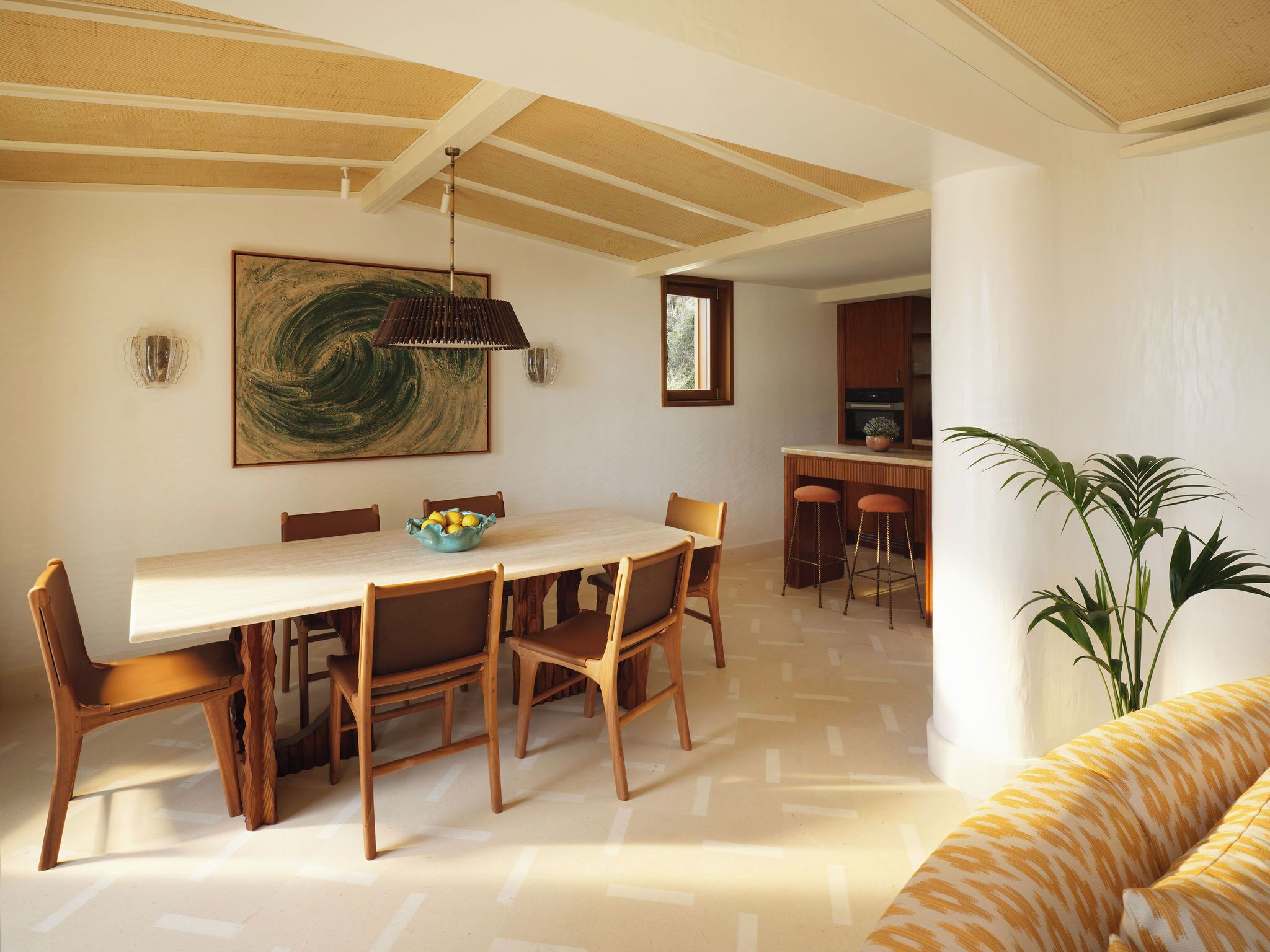 Un espace repas avec une grande table, des chaises, un luminaire suspendu, un tableau et une cuisine en arrière-plan.