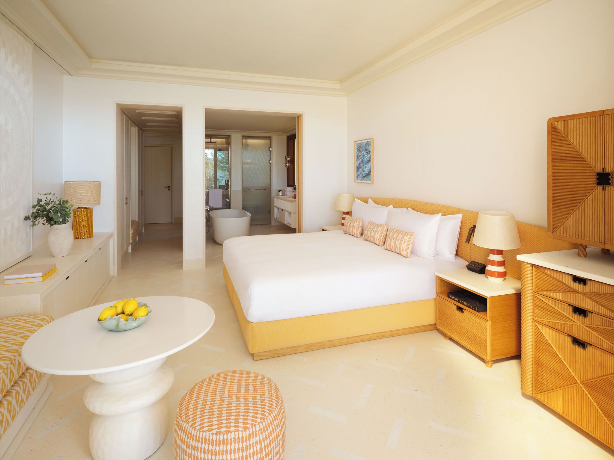 A hotel room with a large bed, bedside tables, a round table with a bowl of fruit, a wooden cabinet, and a bathroom visible through an open door.