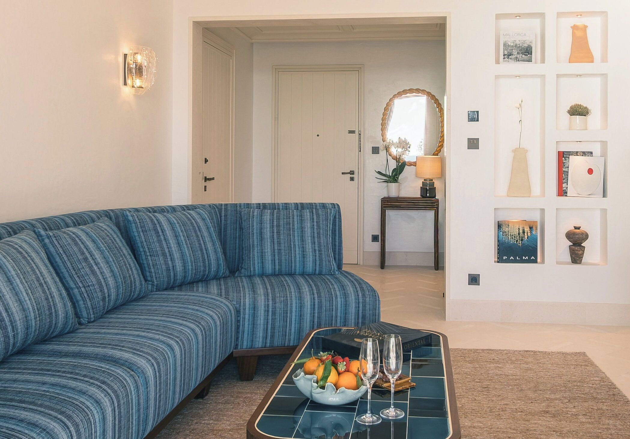 Un salon avec un canapé bleu, une table basse sur laquelle se trouvent des fruits et des verres à vin, un miroir et un mur orné de tableaux et d’un vase.