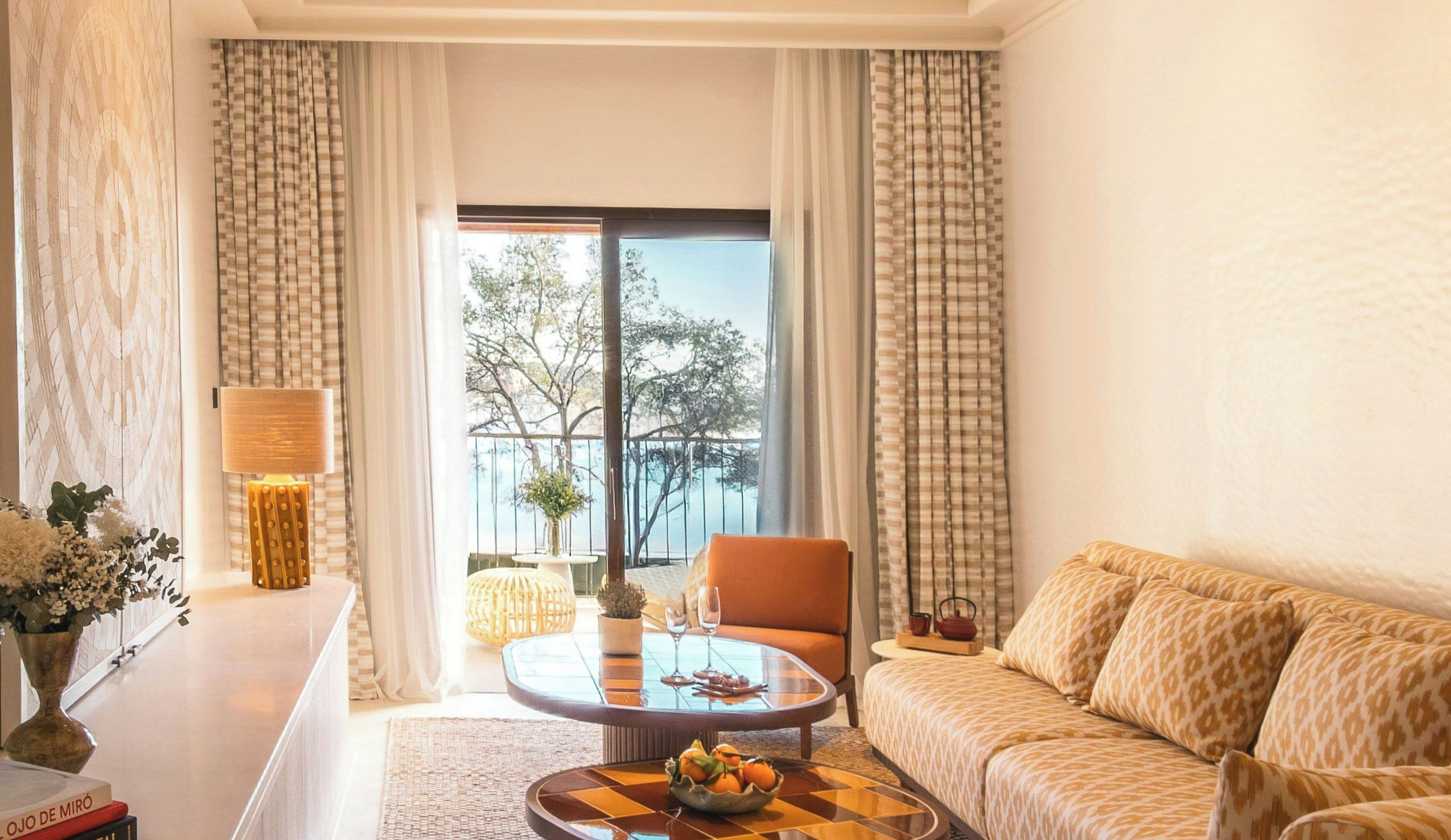 A living room with a sofa, a round coffee table with fruit, a lamp, and a view of a balcony with a railing.