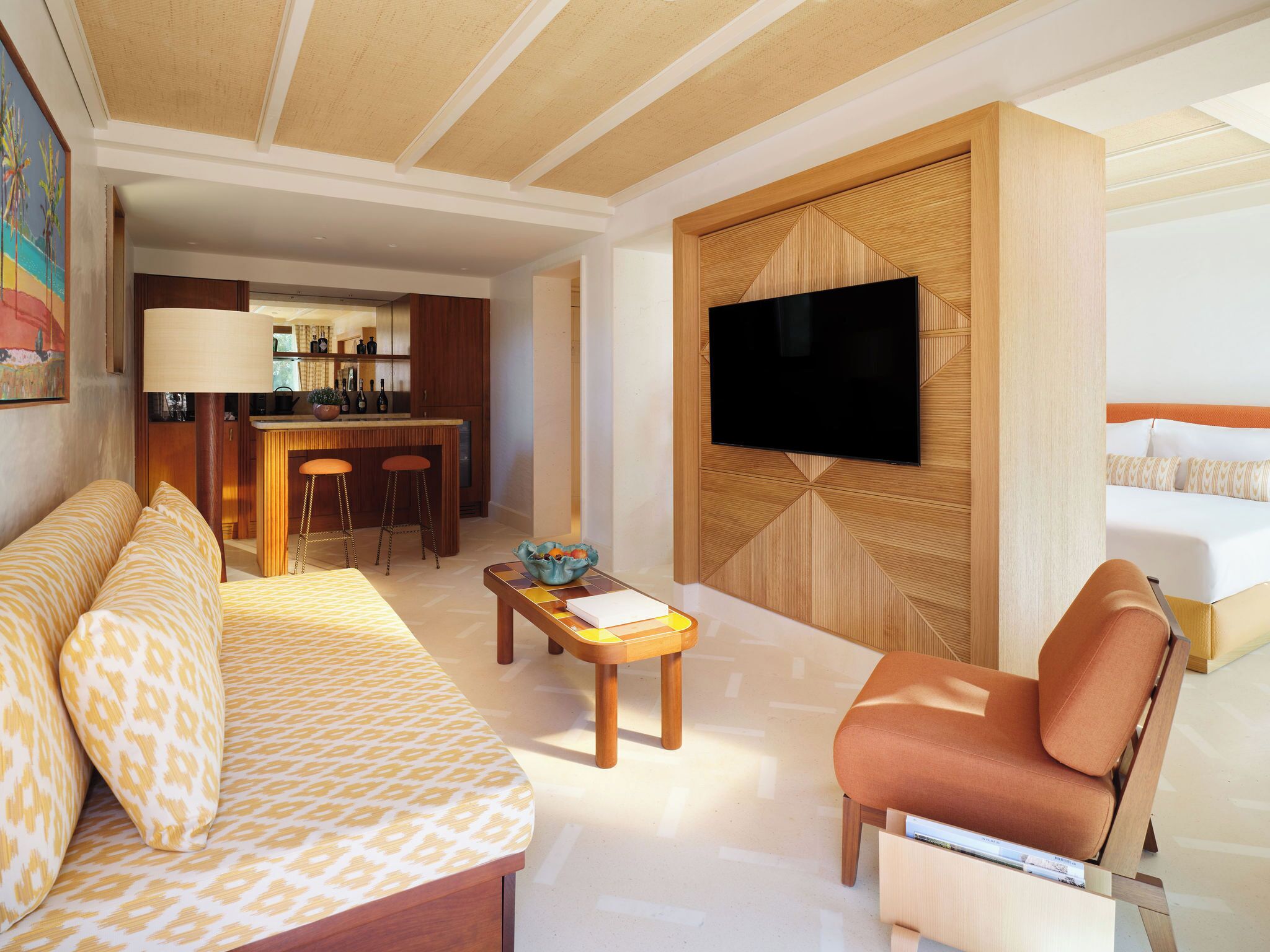 A hotel room with a sofa, a chair, a TV mounted on a wooden panel, a small table with a bowl, and a kitchen area with a bar and stools.