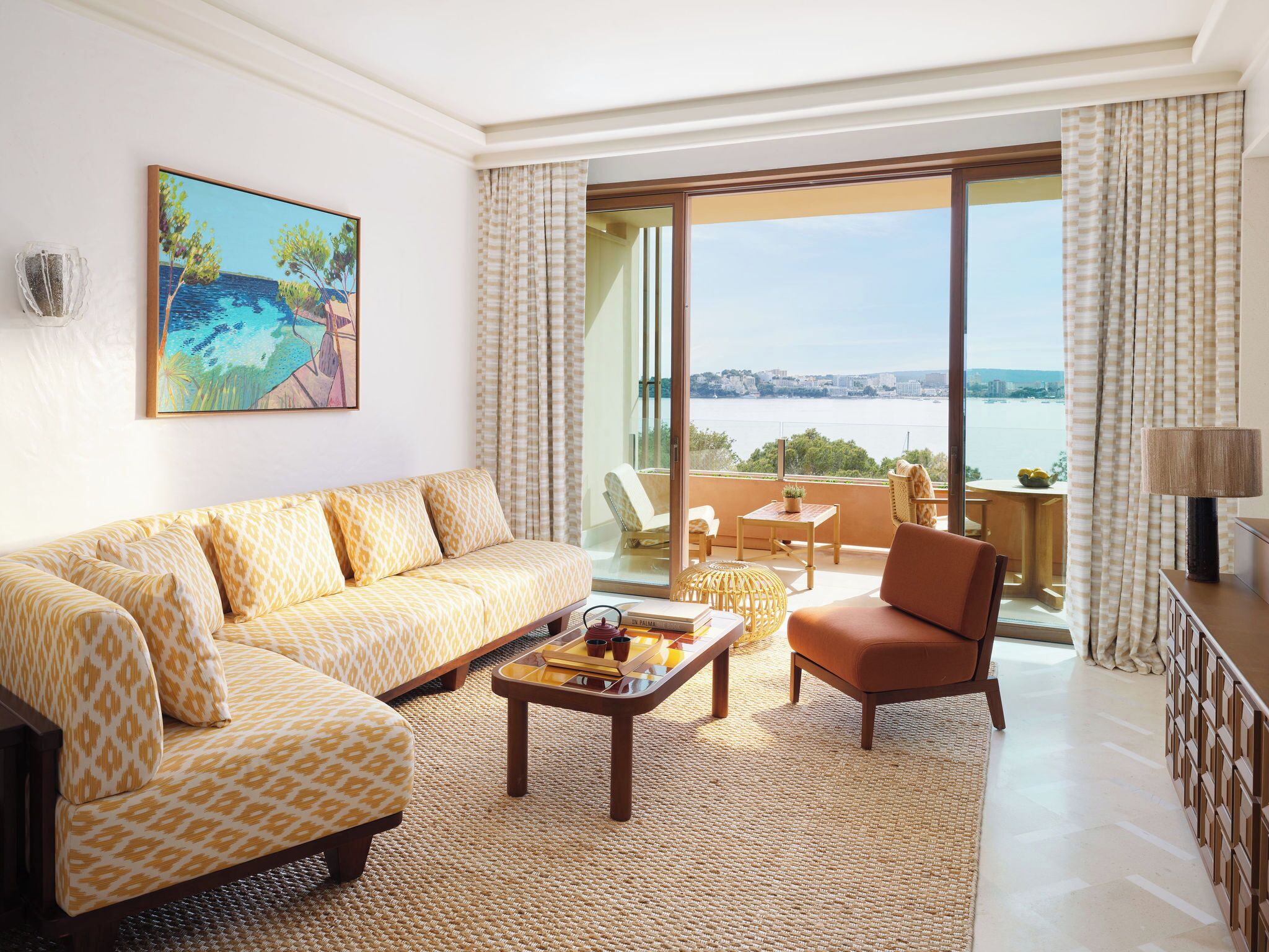 A spacious living room with a sofa, armchair, and coffee table, a painting on the wall, and a balcony with a view of the sea.