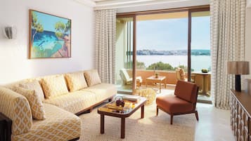 A spacious living room with a sofa, armchair, and coffee table, a painting on the wall, and a balcony with a view of the sea.