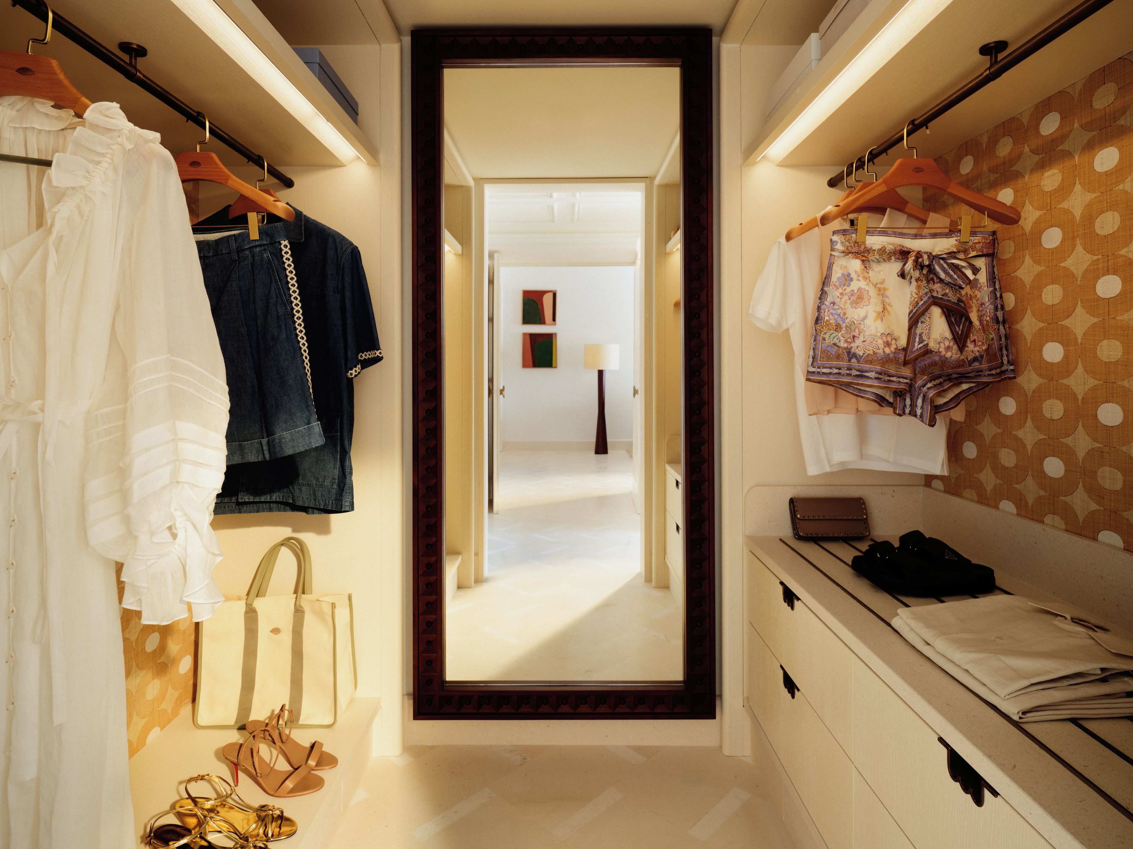 A hallway with clothing racks on both sides, leading to a room with a bed and a nightstand.