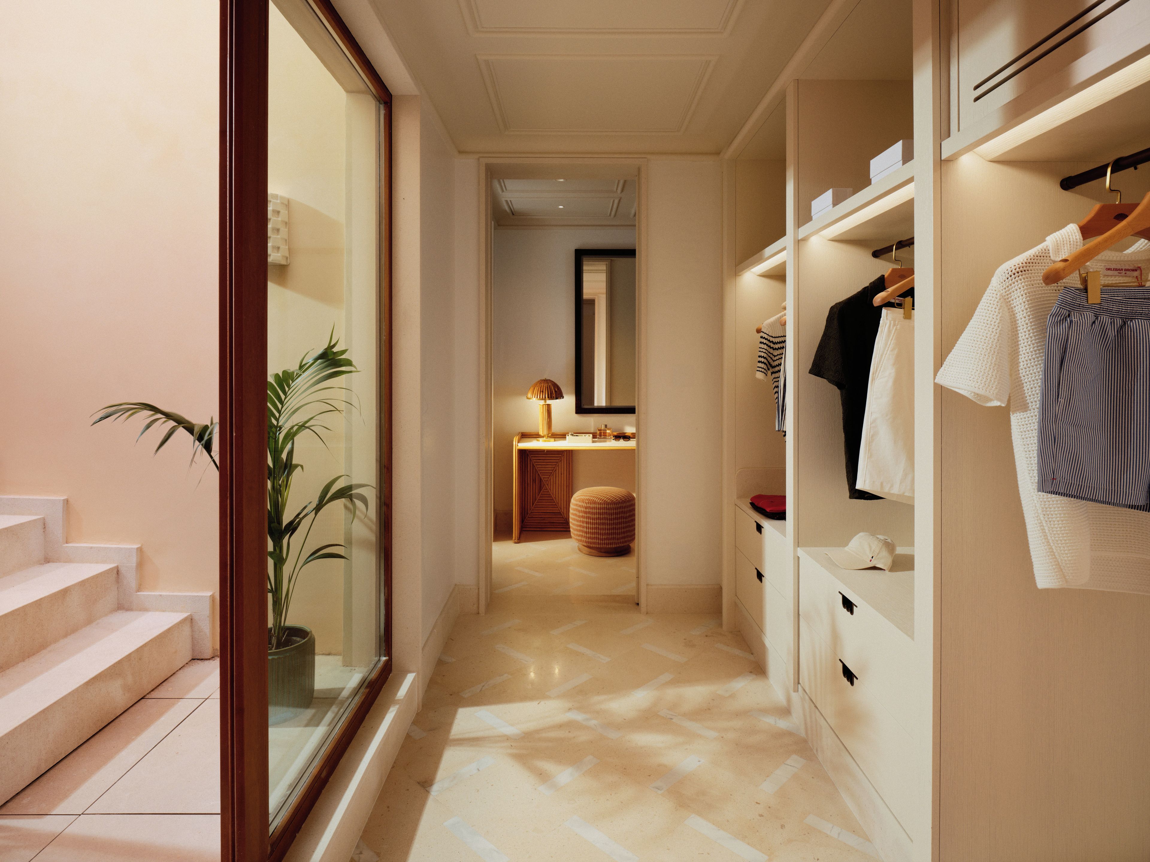 A hallway with a staircase, a plant, a lamp, and a closet with hanging clothes.