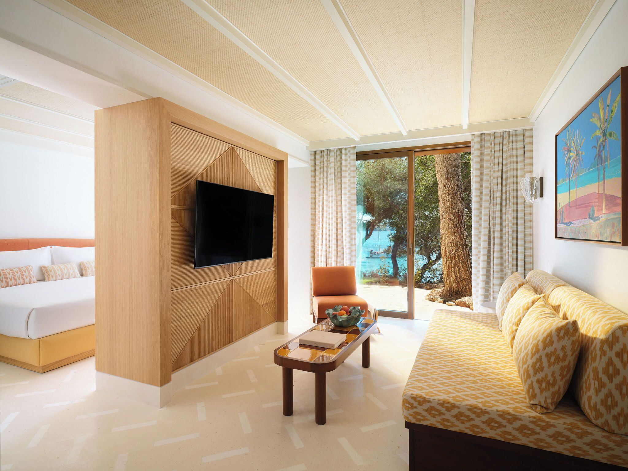 A modern hotel room with a large TV mounted on a wooden panel, a sofa, a coffee table, and a view of the outdoors.