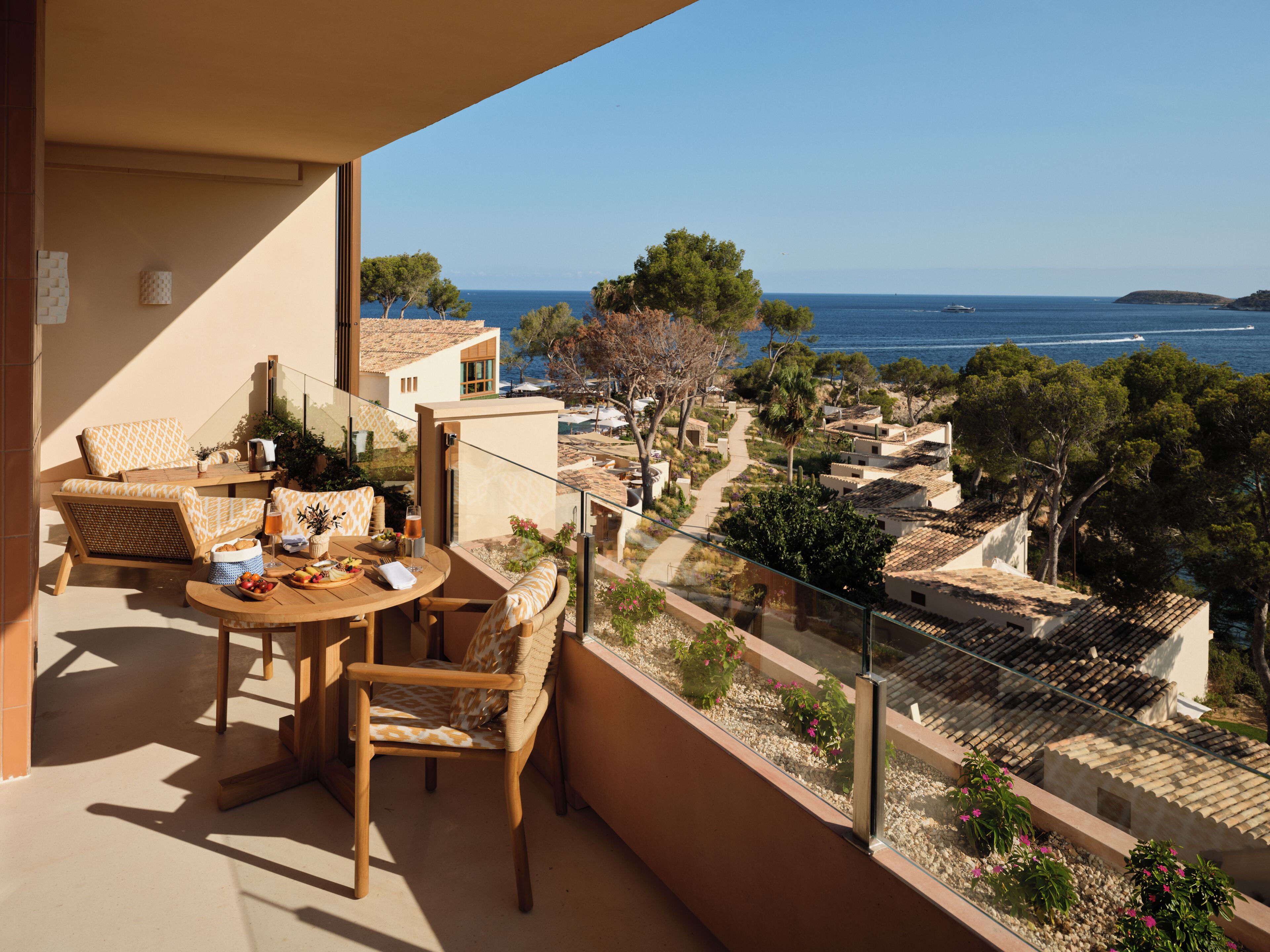 Ein Balkon mit Tisch und Stühlen, der Blick auf ein Küstendorf mit Gebäuden und dem Meer freigibt.