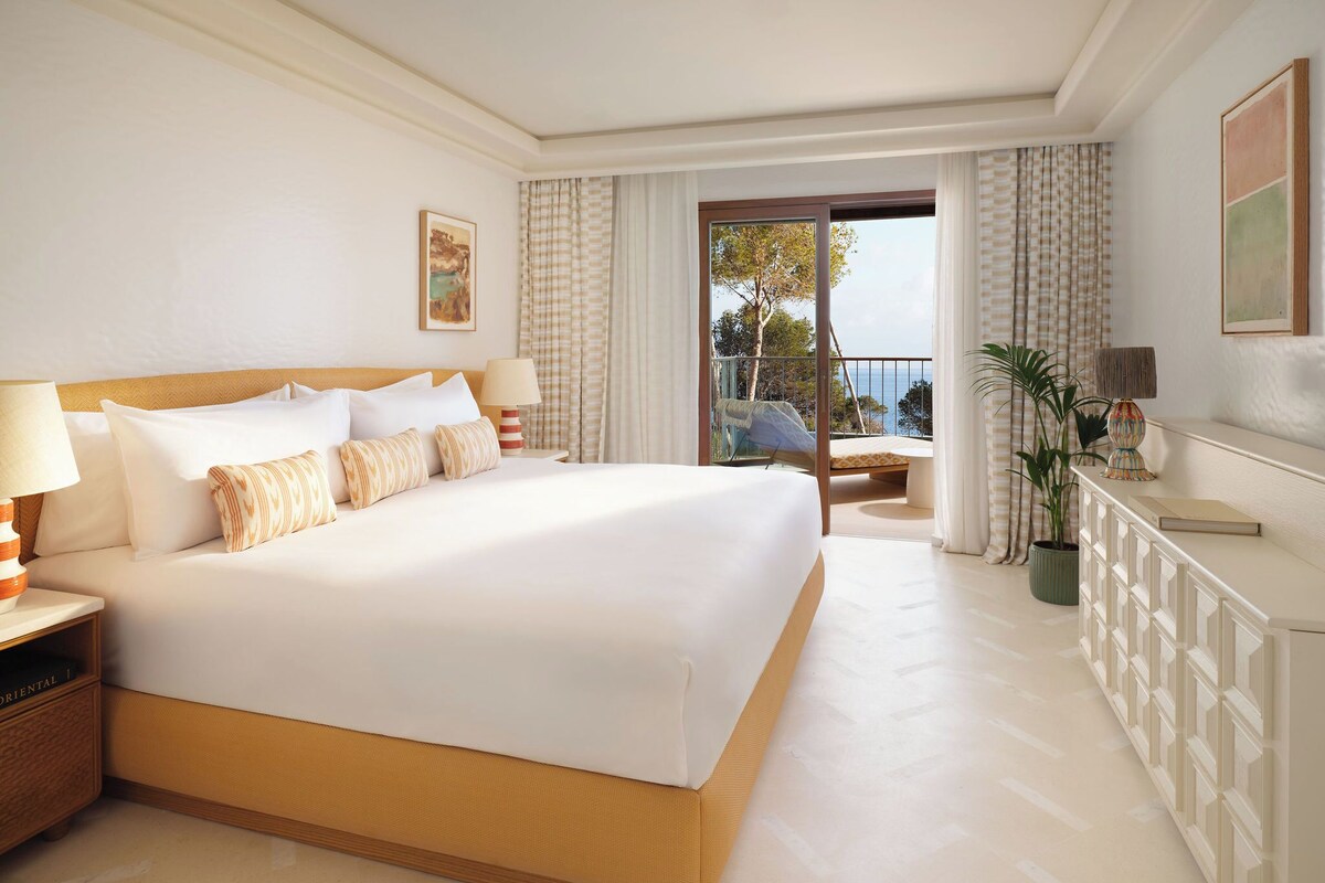 A hotel room with a large bed, a nightstand, a desk, a chair, and a view of the outdoors.
