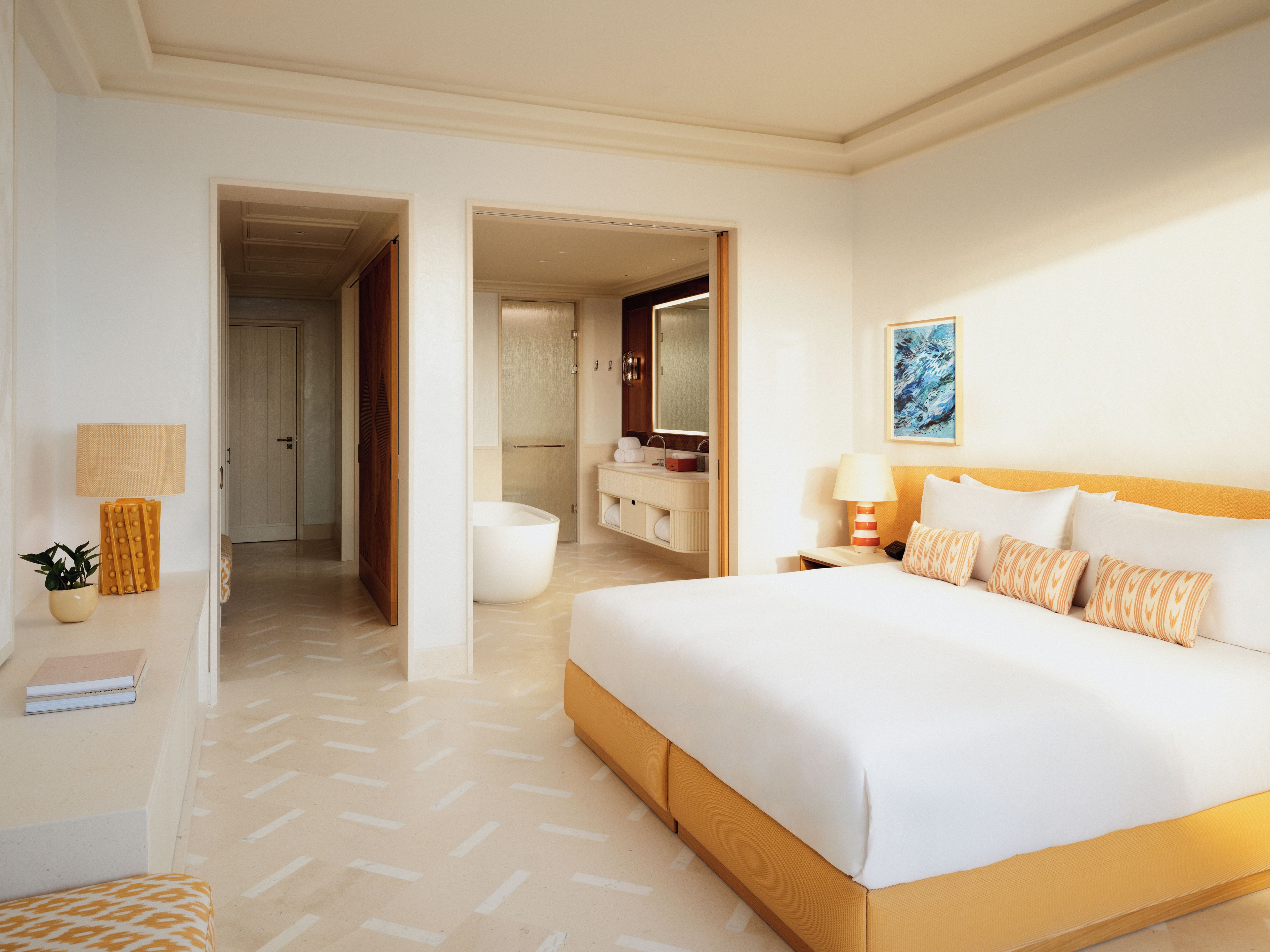 A hotel room with a large bed, a bedside table with a lamp, a bathroom with a bathtub, and a hallway leading to other rooms.