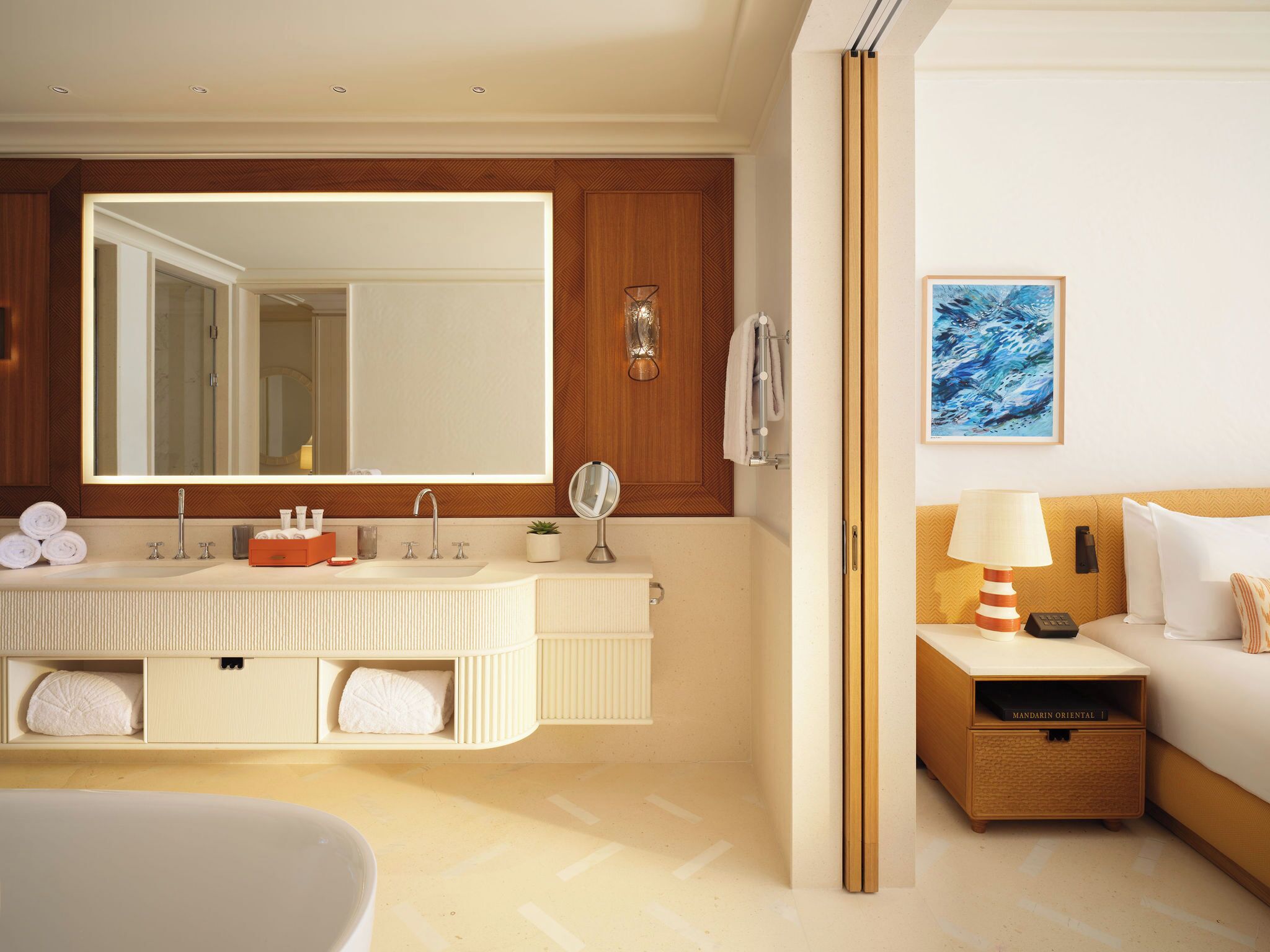 A hotel bathroom with a large mirror, dual sinks, a bathtub, and a bedside table with a lamp.