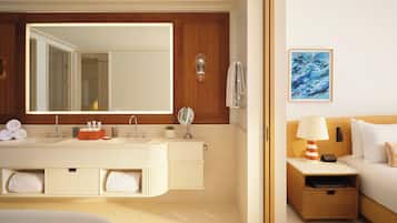 A hotel bathroom with a large mirror, dual sinks, a bathtub, and a bedside table with a lamp.