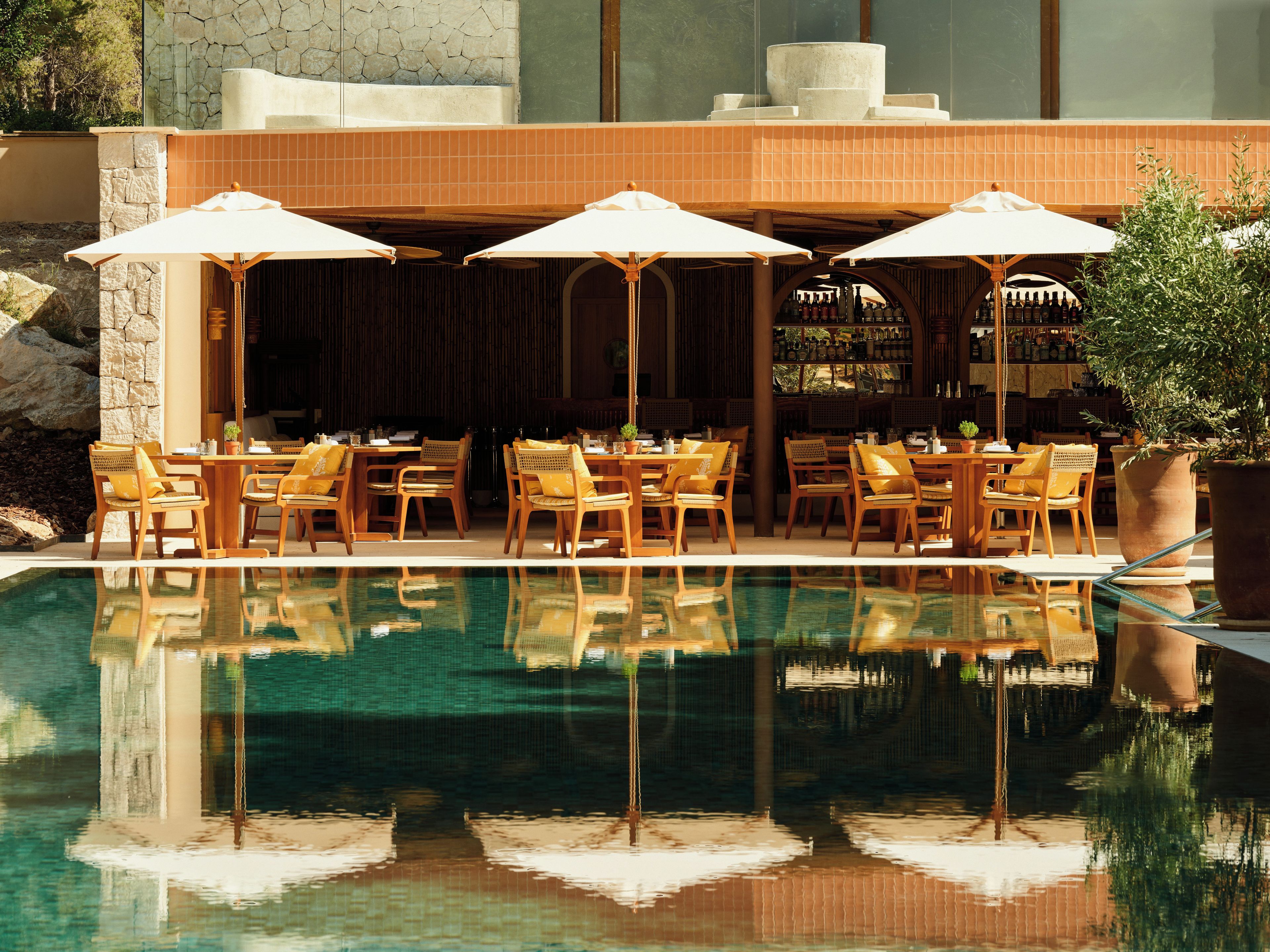 A poolside area with wooden chairs and umbrellas, a stone wall, and a bar area with bottles.