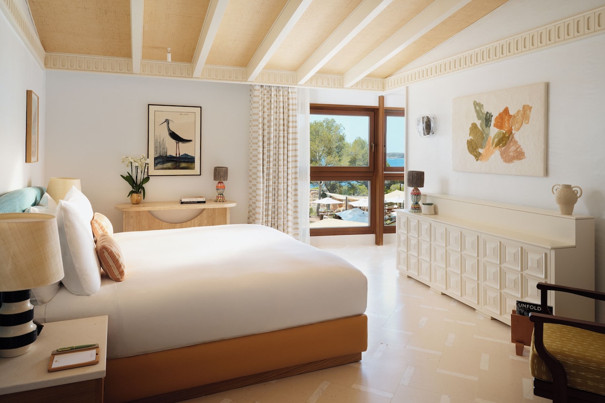 Une chambre spacieuse avec un grand lit, une table de chevet avec des lampes, une commode et une vue sur l’extérieur, ainsi qu’une commode blanche ornée d’un vase.