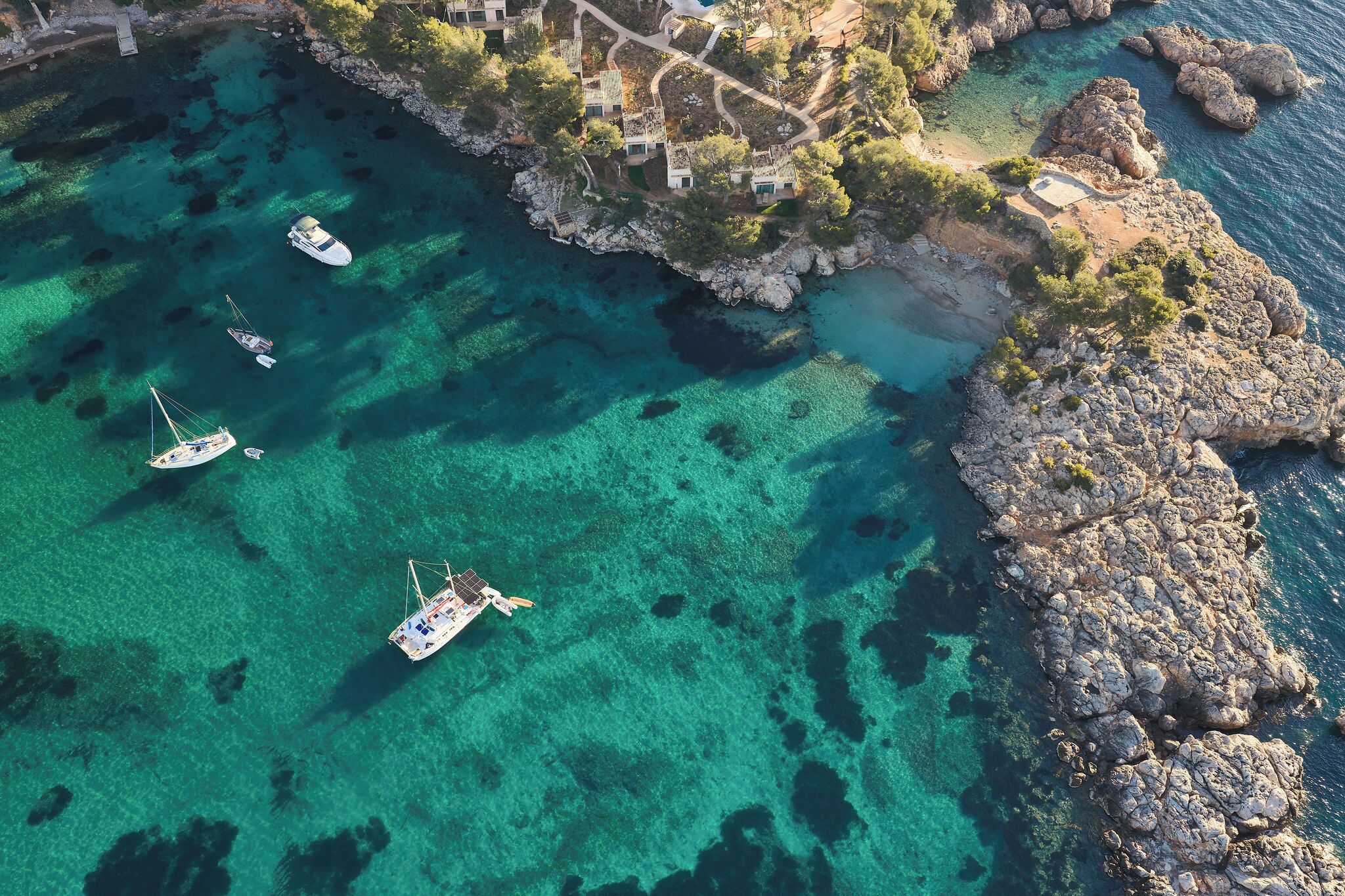 An aerial view of a coastal area with clear turquoise waters, rocky terrain, and several boats anchored.