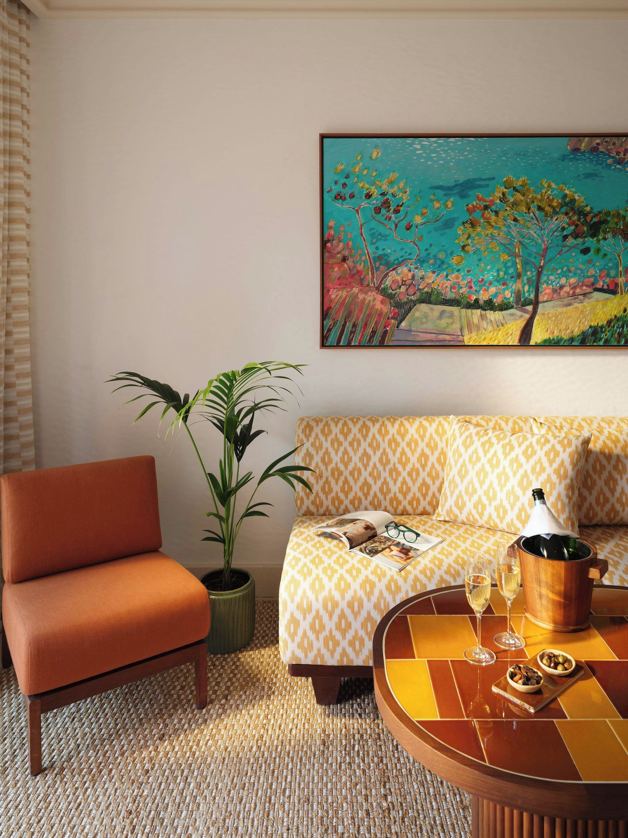 A room with a sofa, a chair, a table with a bottle and glasses, a plant, and a painting on the wall.
